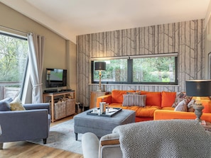 Ein Wohnzimmer mit Sofa, Sesseln, einem Hocker, einem Fernseher und einem Fenster mit Blick auf Grünflächen.