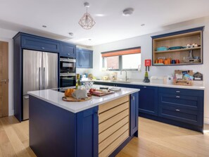 Eine moderne Küche mit blauen Schränken, einem Edelstahlkühlschrank, einem Backofen, einer großen Kochinsel mit Schneidebrett und verschiedenen Küchenutensilien auf der Arbeitsfläche.