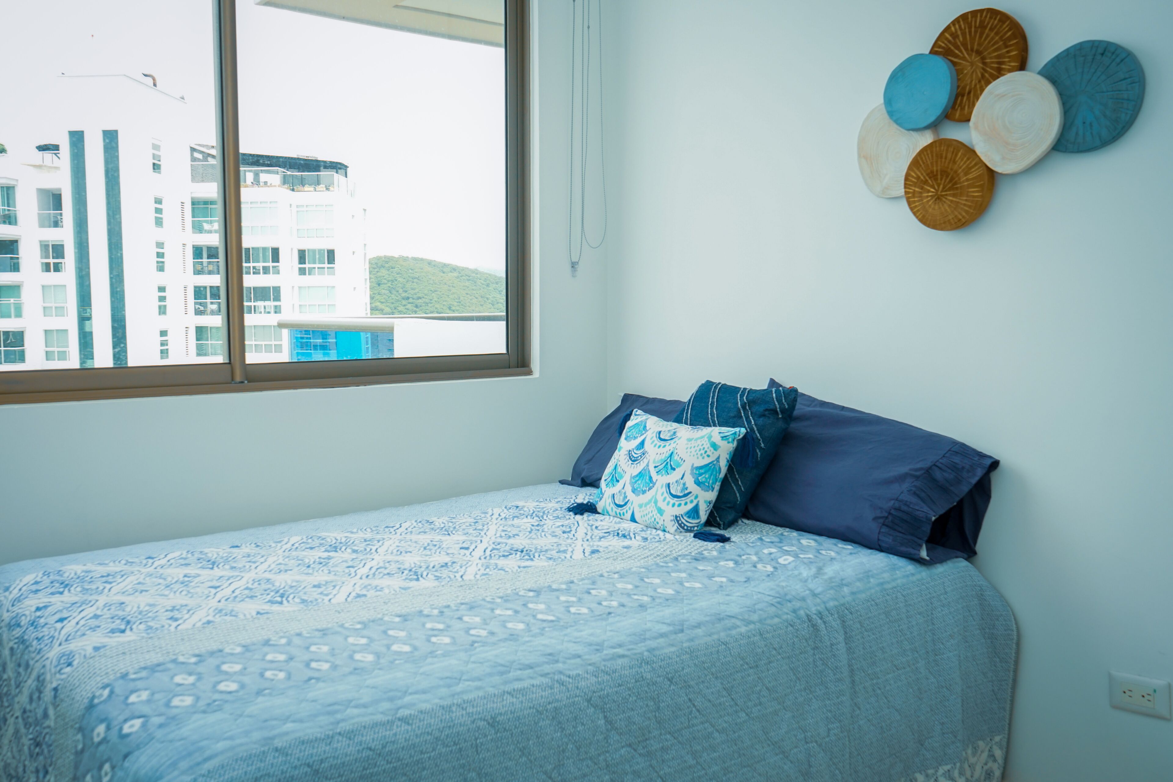 Een slaapkamer met een bed, kussens, een decoratie aan de muur en een raam met uitzicht op gebouwen.