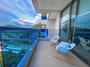 A balcony with a glass railing, two rocking chairs, and a view of a cityscape and mountains. - Luxury Resort Condo in Santa Marta Walk to Beach (Santa Marta)