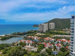 A coastal area with a beach, a small island, and a hill. There are buildings with red roofs and a tall white building. - Luxury Resort Condo in Santa Marta Walk to Beach (Santa Marta)