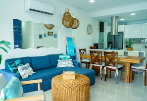 A modern living room with a blue sofa, wooden dining table, wicker ottoman, and kitchen area with white cabinets. - Luxury Resort Condo in Santa Marta Walk to Beach (Santa Marta)
