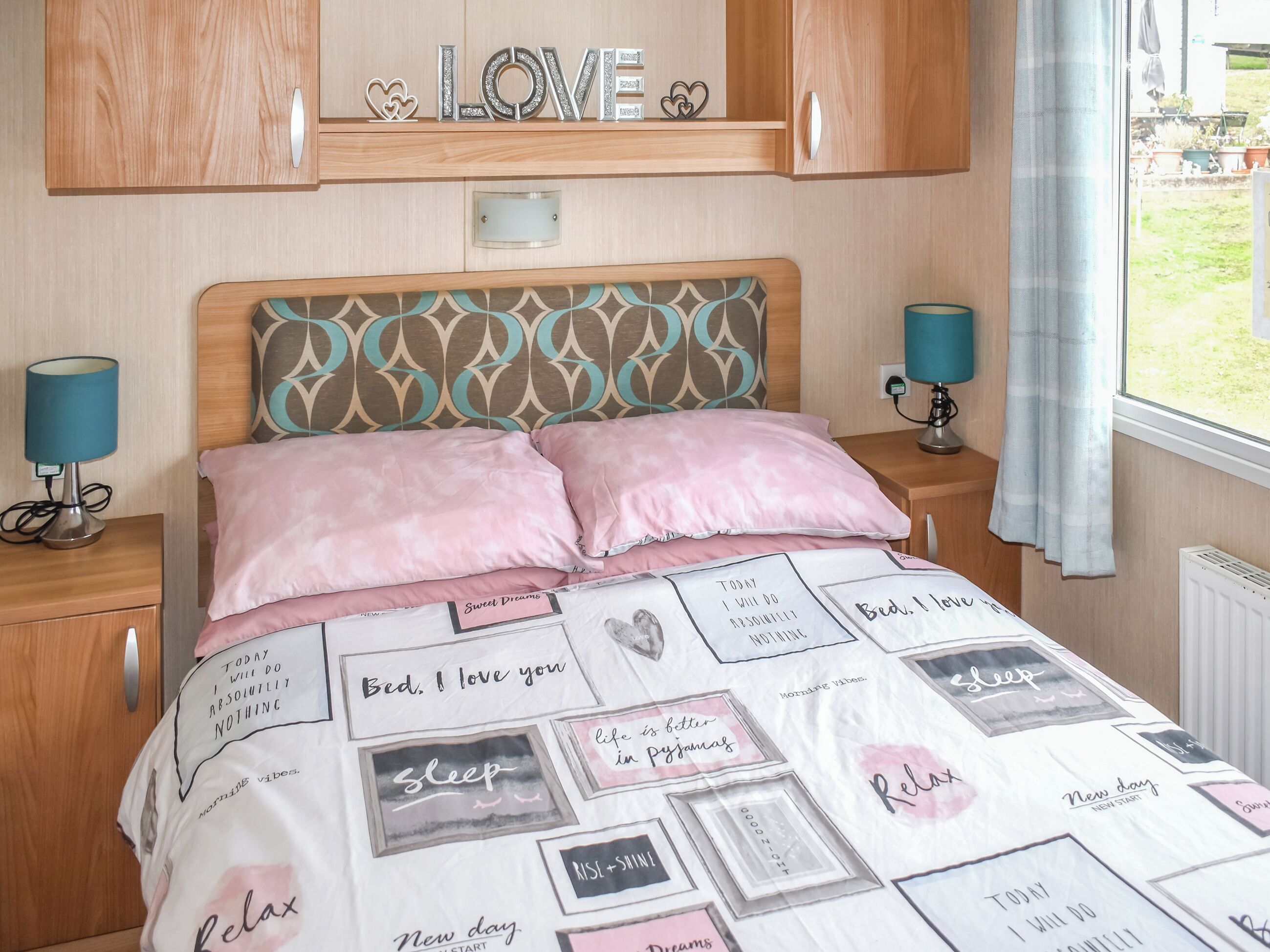 A bedroom with a bed, two bedside lamps, a headboard with a decorative panel, a window with curtains, and a wall-mounted shelf with a 'LOVE' sign.