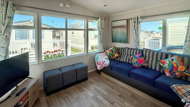 A modern living room with a sofa, a bench, a TV, and large windows offering a view of houses. - 3 bedroom accommodation in Towyn (Towyn)