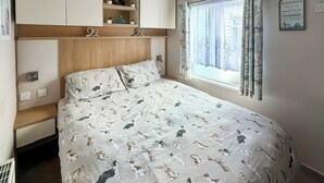 A bedroom with a bed, built-in shelving, a window with curtains, and a wall-mounted sign. - 3 bedroom accommodation in Towyn (Towyn)