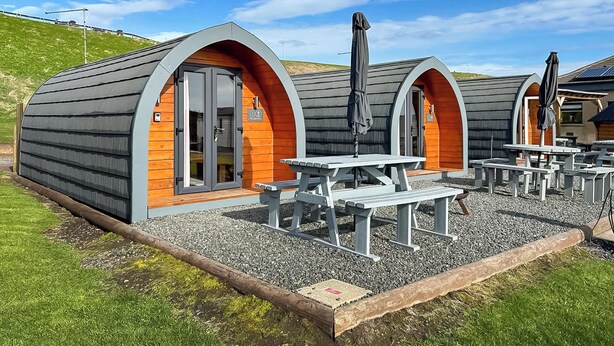 A row of small, semi-circular huts with wooden walls and grey roofs, each with a door and a window, and outdoor seating areas with tables and benches. - Relax in our stunning seaside glamping pods in Peterhead. (Banff)