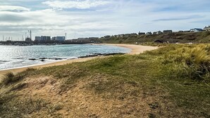 A coastal landscape with a sandy beach, grassy dunes, and a distant cityscape with buildings and cranes. - Relax in our stunning seaside glamping pods in Peterhead. (Banff)