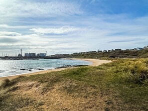 Un paisaje costero con una playa arenosa, dunas cubiertas de hierba y una ciudadela distante con edificios y grúas.