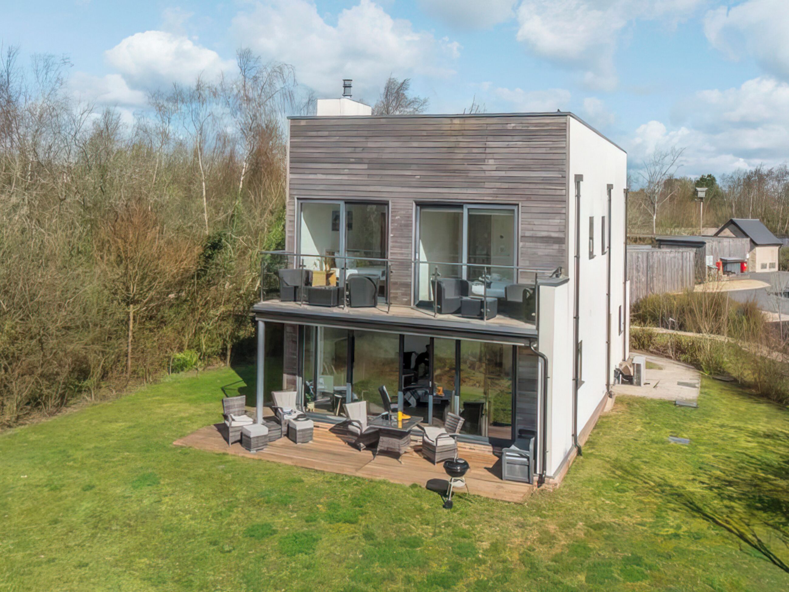 Una casa moderna de dos pisos con balcón, ventanas amplias y una terraza con mobiliario exterior.