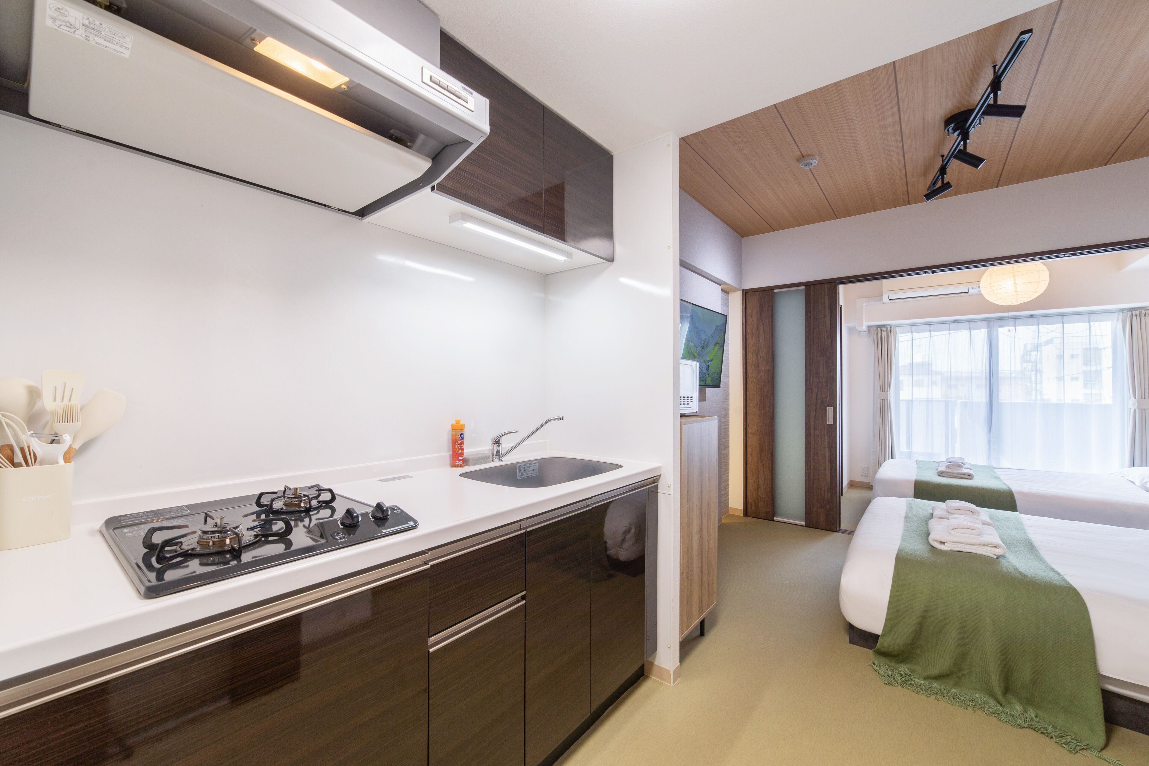 A modern kitchen with a gas stove, sink, and white countertops. There is a bedroom with a bed and a green throw blanket.