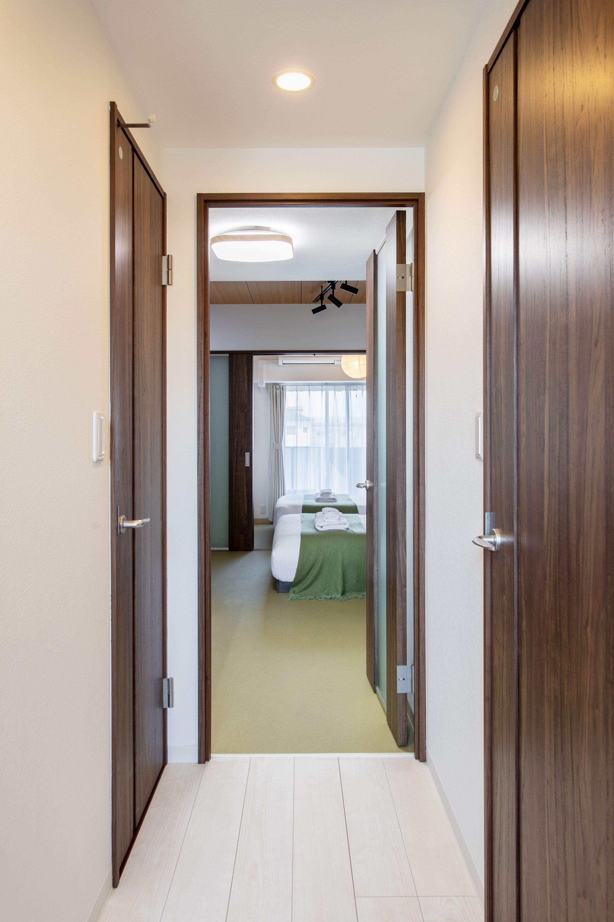 A hotel corridor leading to a room with a bed, a bedside table, and a window with curtains.
