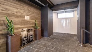 Zona de recepción con pared de madera, una planta en maceta, un perchero metálico para paraguas y suelo de baldosas.