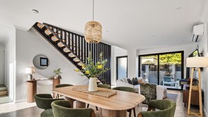 Un comedor moderno con una mesa de madera, sillas tapizadas en verde, una lámpara colgante y una escalera con barandillas negras.