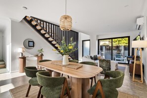 A modern dining area with a wooden table, green upholstered chairs, a hanging light fixture, and a staircase with black railings. - Stone River Villa 3 | Relaxed Stay | Heated Pool (Margaret River)