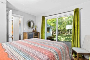Ein Schlafzimmer mit Bett, Sessel, Kommode, Spiegel und einer Glasschiebetür zum Garten.