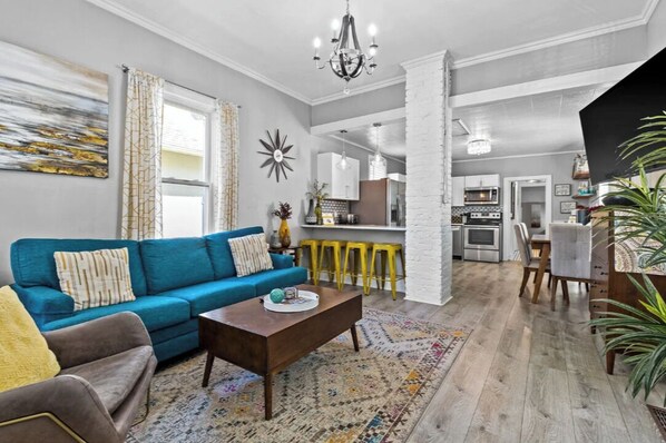 Ein Wohnzimmer mit einem blauen Sofa, einem Couchtisch und einem gemusterten Teppich. Im Hintergrund ist eine Küche mit einer Bar erkennbar.