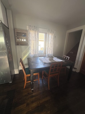 A dining area with a wooden table, chairs, a window with curtains, a refrigerator, and a wall-mounted shelf.