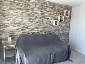 A room with a stone-patterned wall, a covered grey sofa, a side table, and a white cabinet with shelves. - Capitole, seafront, studio with sleeping alcove for 3 people, 26m2, balcony with partial sea view, air conditioning, private parking (Fréjus)