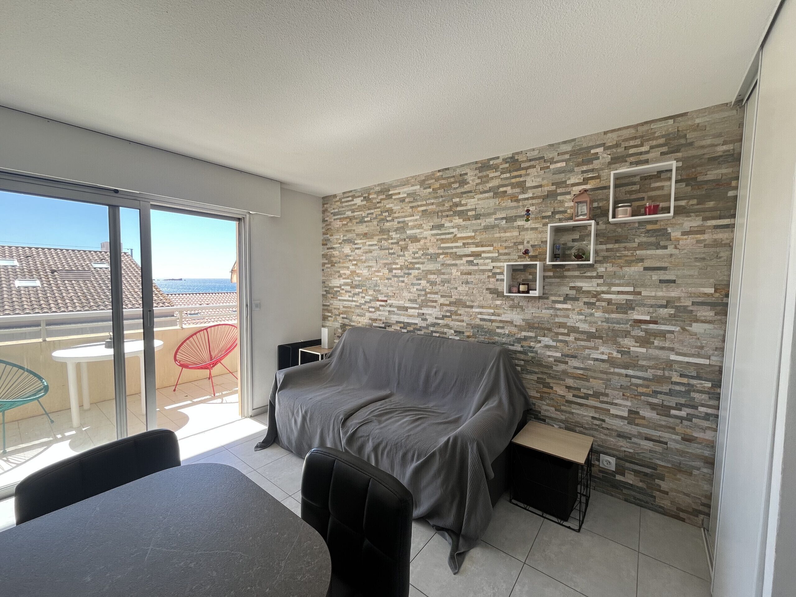 Un balcon avec vue sur les immeubles et la mer, un canapé couvert, une petite table et une chaise.