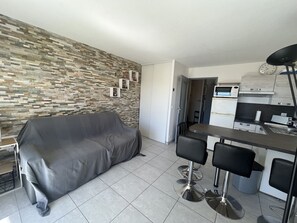 A living room with a covered sofa, a dining area with bar stools, a kitchen with modern appliances, and a patterned wall. - Capitole, seafront, studio with sleeping alcove for 3 people, 26m2, balcony with partial sea view, air conditioning, private parking (Fréjus)