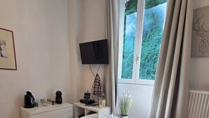 A room with a dining table set for two, a TV mounted on the wall, a window with curtains, a radiator, and a bed with a white cover. - Charming studio in the heart of uriage (saint martin d'uriage)