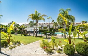 A pool area with palm trees, a pathway, and a building with a tiled roof in the background. - Solmare, Coastal Comfort in Estepona (Estepona)