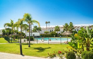 A pool area with a fenced pool, palm trees, and a building with a tiled roof in the background. - Solmare, Coastal Comfort in Estepona (Estepona)