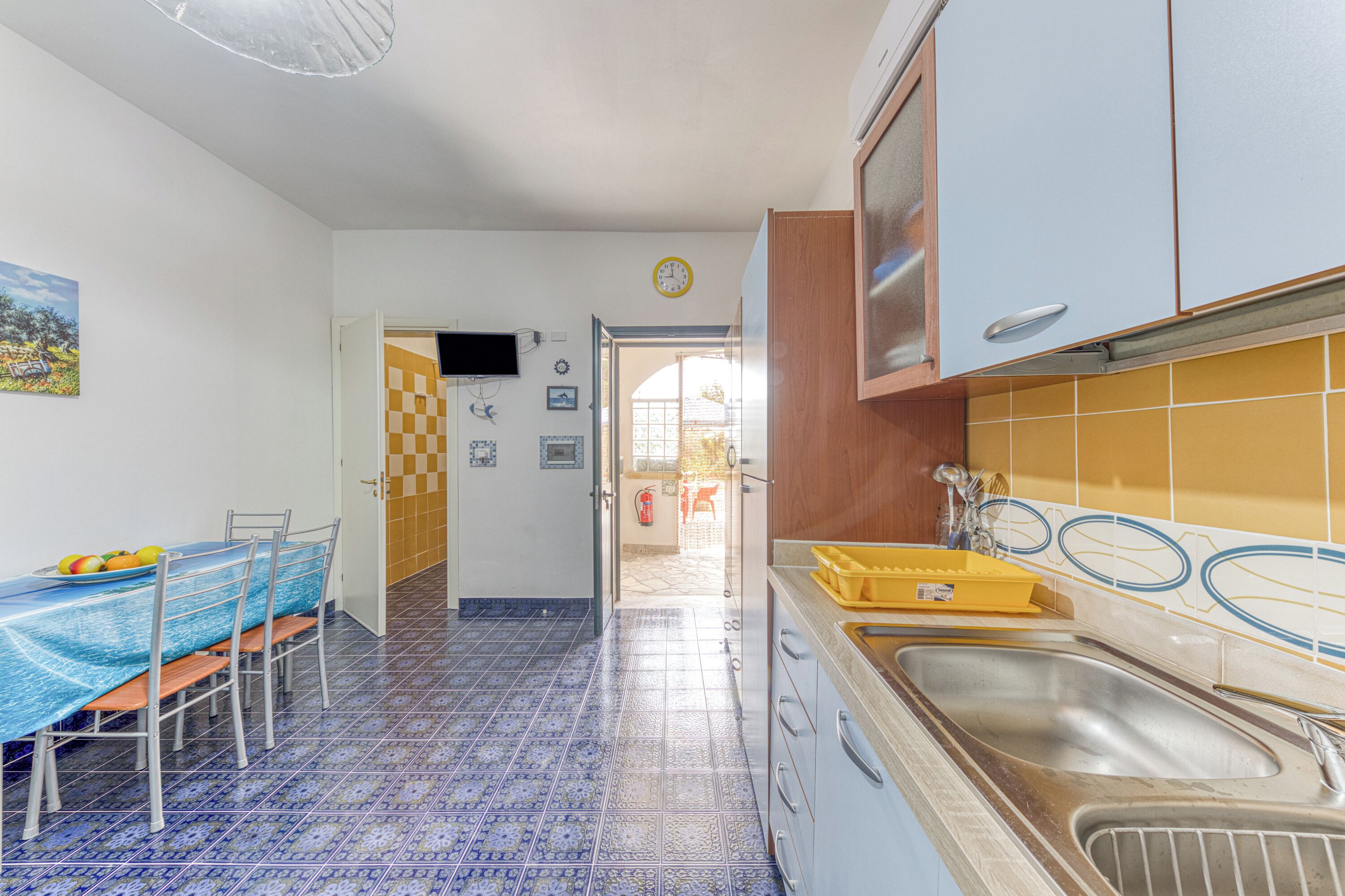 A kitchen with a blue tiled floor, a dining table with chairs, and a sink with a yellow dish rack.
