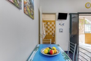 Ein Essbereich mit einem für zwei Personen gedeckten Tisch, einer Obstplatte, einem an der Wand montierten Fernseher und einer Tür zu einem Badezimmer mit Schachbrettmuster.