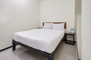 A single bed with a wooden headboard, a small bedside table with a cup and water bottles, and a plain white wall.