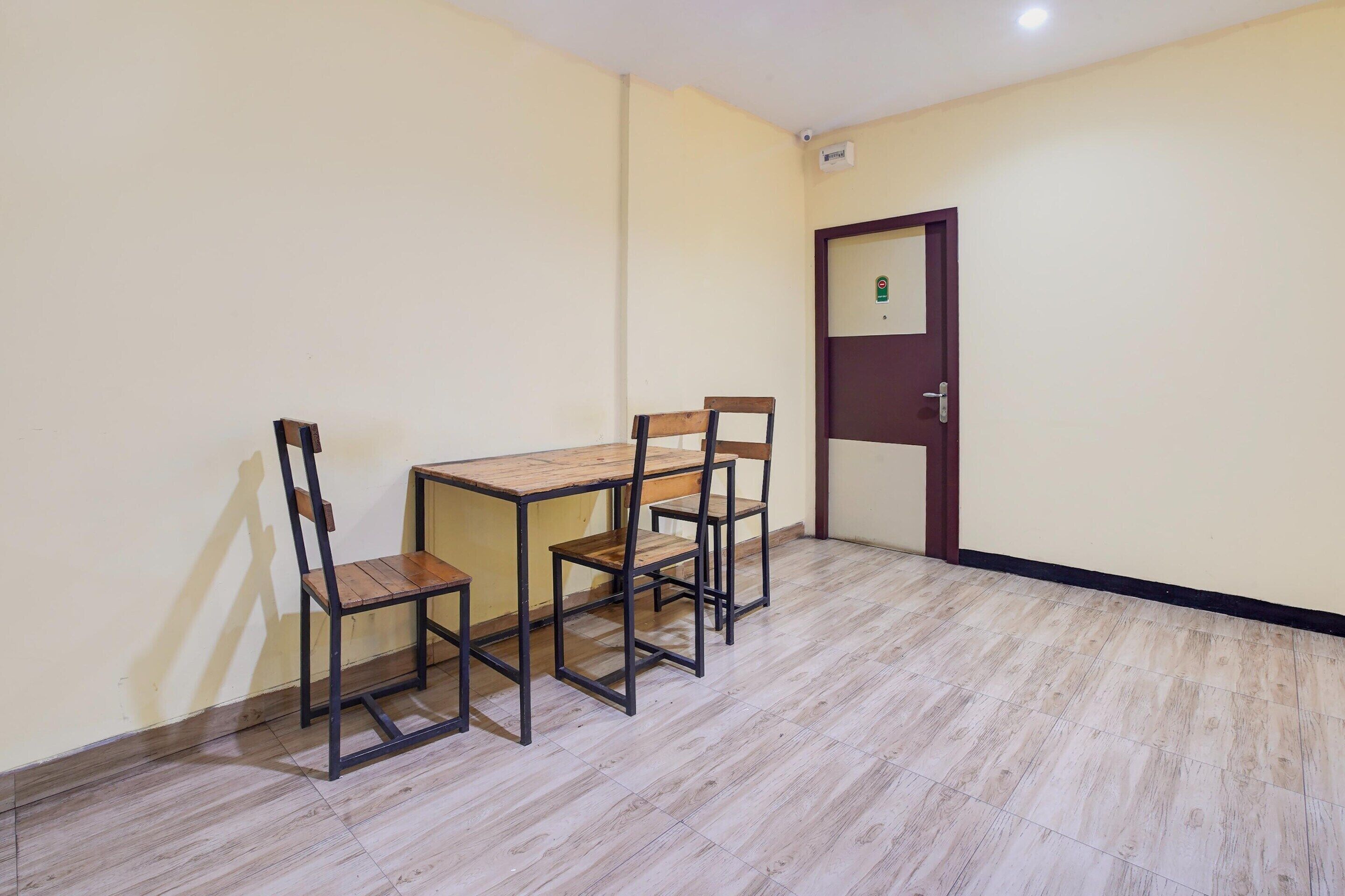 A room with a wooden table and four chairs, a door with a security camera, and a tiled floor.