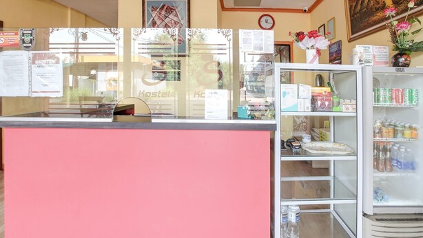 Balcão de recepção com bancada rosa, vitrine de vidro com diversos itens e uma geladeira abastecida com bebidas.