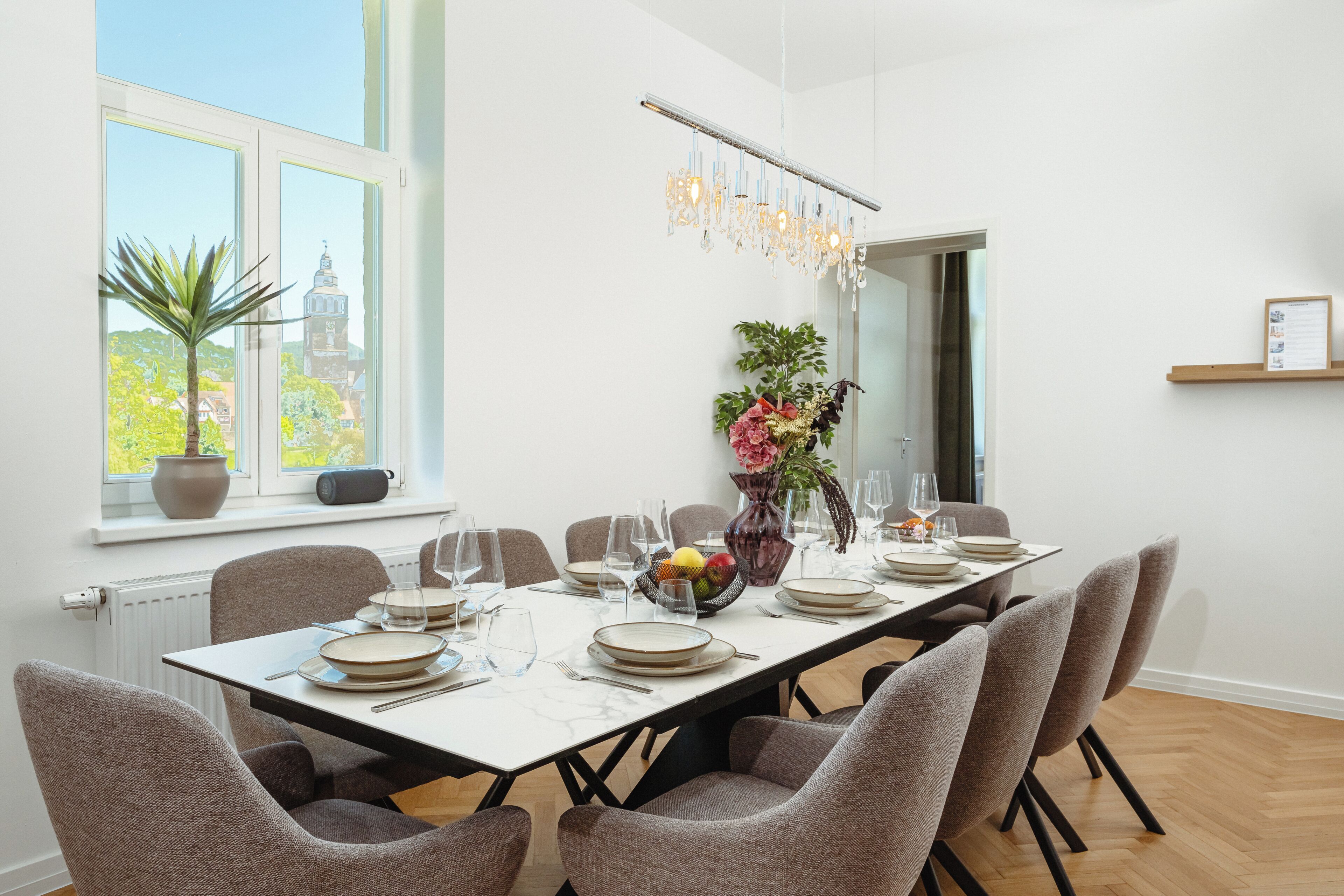 En spiseplads med et langt bord dækket til seks personer, stole, en vase med blomster, en skål med frugt og en hængende lysekrone.