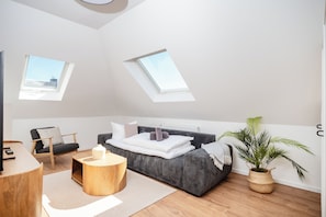 A modern bedroom with a skylight, a sofa, a chair, a round wooden side table, and a potted plant.