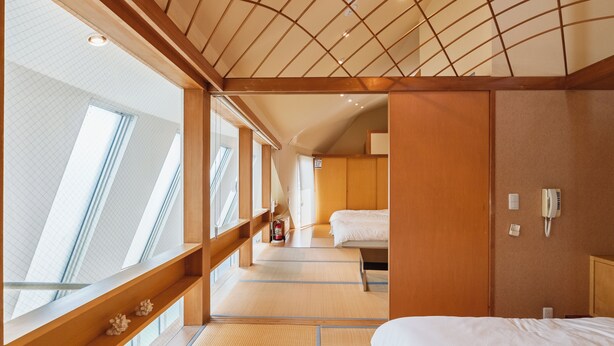 Una habitación de hotel con suelo de tatami, techo de madera, cama, mesita de noche y teléfono montado en la pared, y una ventana con vistas.