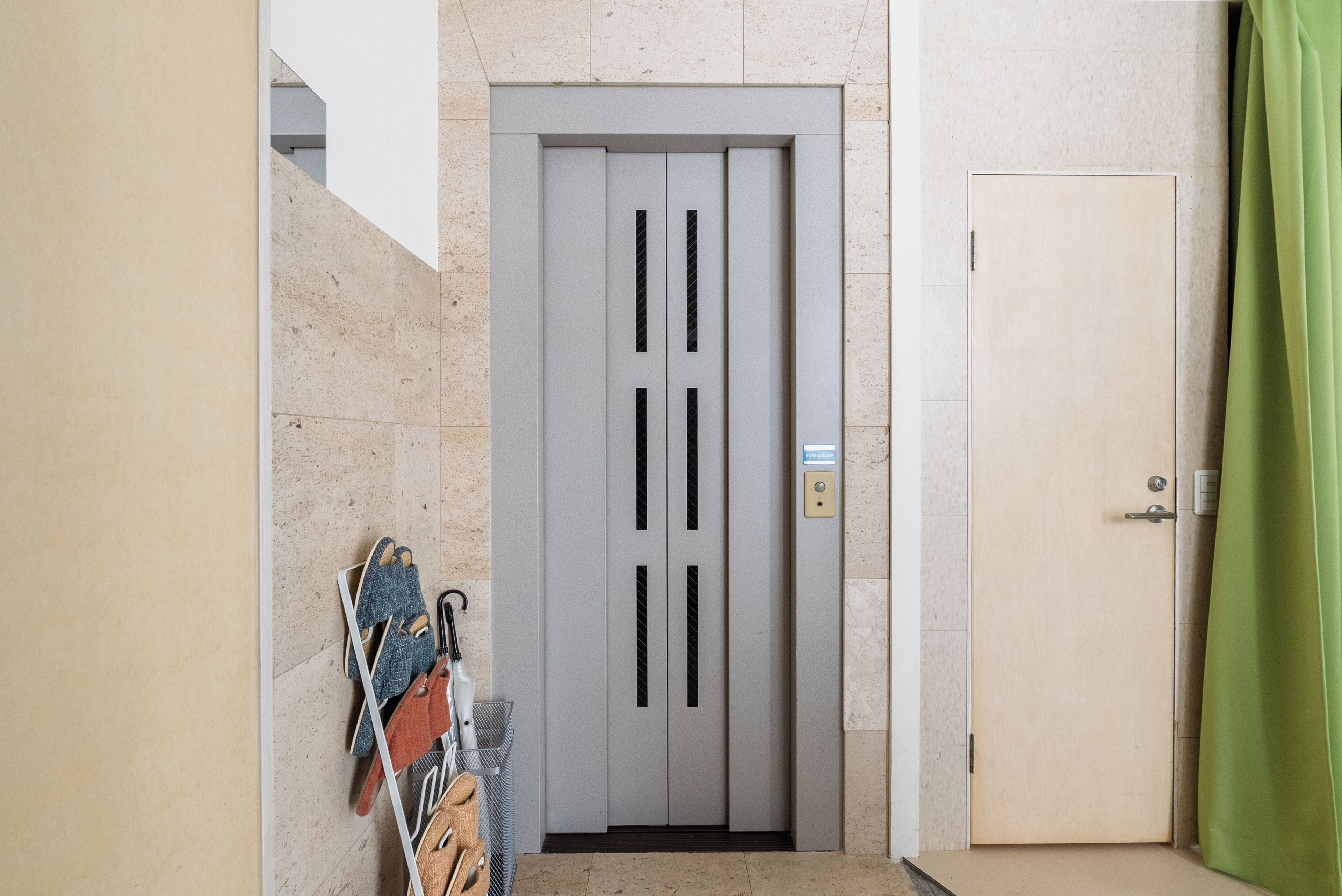 Un pasillo con un ascensor gris, una puerta con manija y un perchero metálico con zapatos.