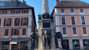 Et monument med statuer af elefanter og en figur, omgivet af bygninger med mange vinduer og et belagt område foran.
