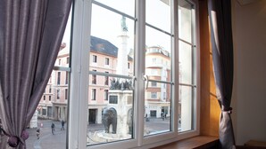 Et vindue med udsigt til et torv, en statue og bygninger i baggrunden.