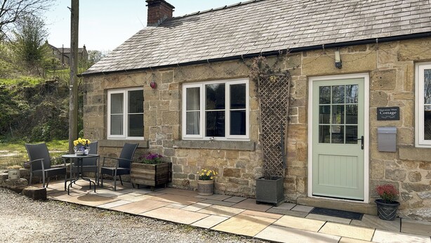 A stone cottage with a green door, two windows, and a small patio with outdoor seating. - Mawstone Mine Cottage (Youlgrave)