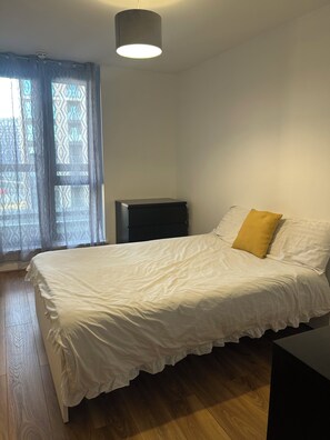 Ein Schlafzimmer mit Bett, Nachttisch, Lampe und einem Fenster mit Vorhängen.