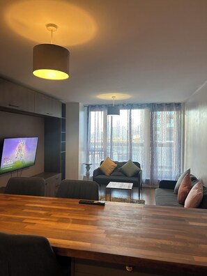 Ein modernes Wohnzimmer mit einem Holzespeisetisch, einer Couch, einem an der Wand befestigten Fernseher und großen Fenstern mit gemusterten Vorhängen.