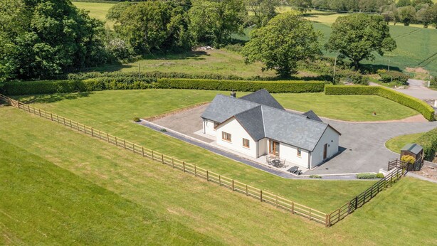 Een huis met een verharde oprit, omringd door een omheind gazon, gelegen in een landelijk gebied met glooiende heuvels en bomen op de achtergrond.