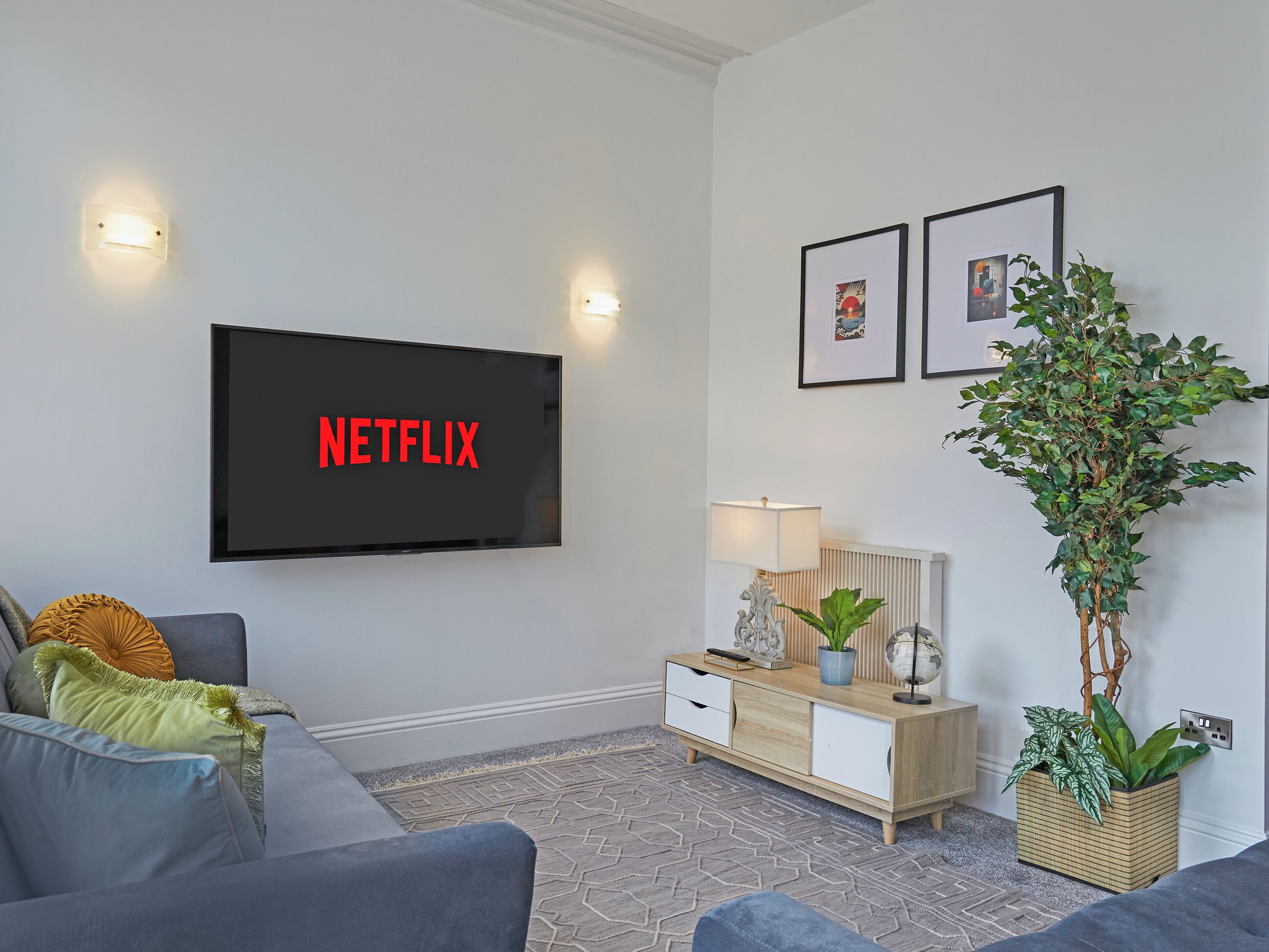 A living room with a flat-screen TV, a sofa, a side table, a lamp, framed pictures, and a potted plant.