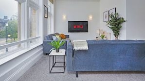 Een moderne woonkamer met een grijze bank, een bijzettafeltje met een plant, een tv aan de muur waarop 'NETFLIX' staat, en grote ramen met uitzicht op de stad.
