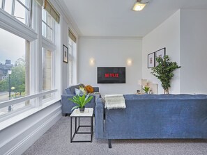 Ein modernes Wohnzimmer mit einem grauen Sofa, einem kleinen Beistelltisch mit einer Pflanze, einem Wand-Fernseher mit der Aufschrift 'NETFLIX' und großen Fenstern mit Blick auf die Stadt.