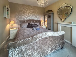 Ein Schlafzimmer mit einem gemusterten Bettbezug, einer Kristallleuchtre, einem herzförmigen Spiegel und einem Schminktisch.