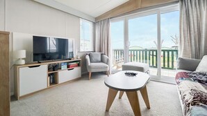 Moderni hotellihuone, jossa on taulutelevisio, sohva, tuoli, pieni pöytä ja parveke, jolta on näkymä.