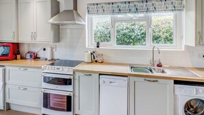 A modern kitchen with white cabinetry, a built-in oven, a dishwasher, a washing machine, and a window with a view of greenery. - Bembridge Coastal Escape (Bembridge)