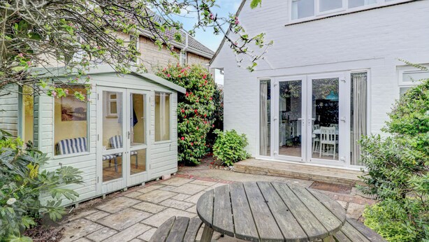 A patio with a wooden table and benches, a small shed, and a white house with a glass door. - Bembridge Coastal Escape (Bembridge)