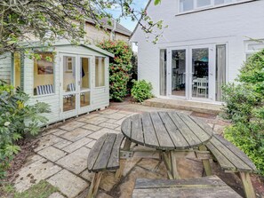 Une terrasse avec une table et des bancs en bois, un petit abri de jardin et une maison blanche avec une porte en verre.